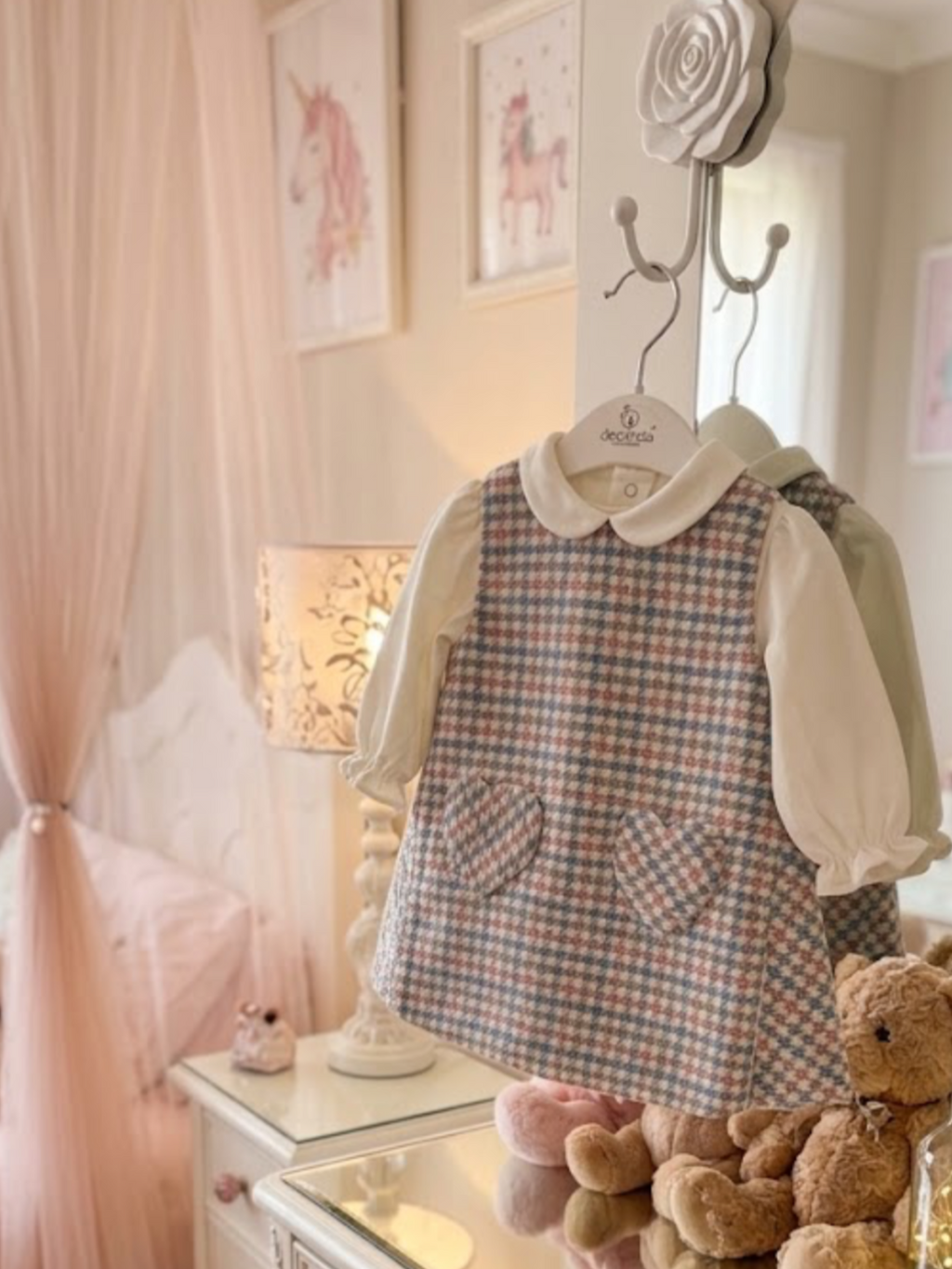 A children's long-sleeve dress with a blue, pink, and beige gingham pinafore-style front panel over a cream puff-sleeve blouse, displayed on a hanger on a wall rack in a soft pink bedroom setting with teddy bears and framed unicorn art.